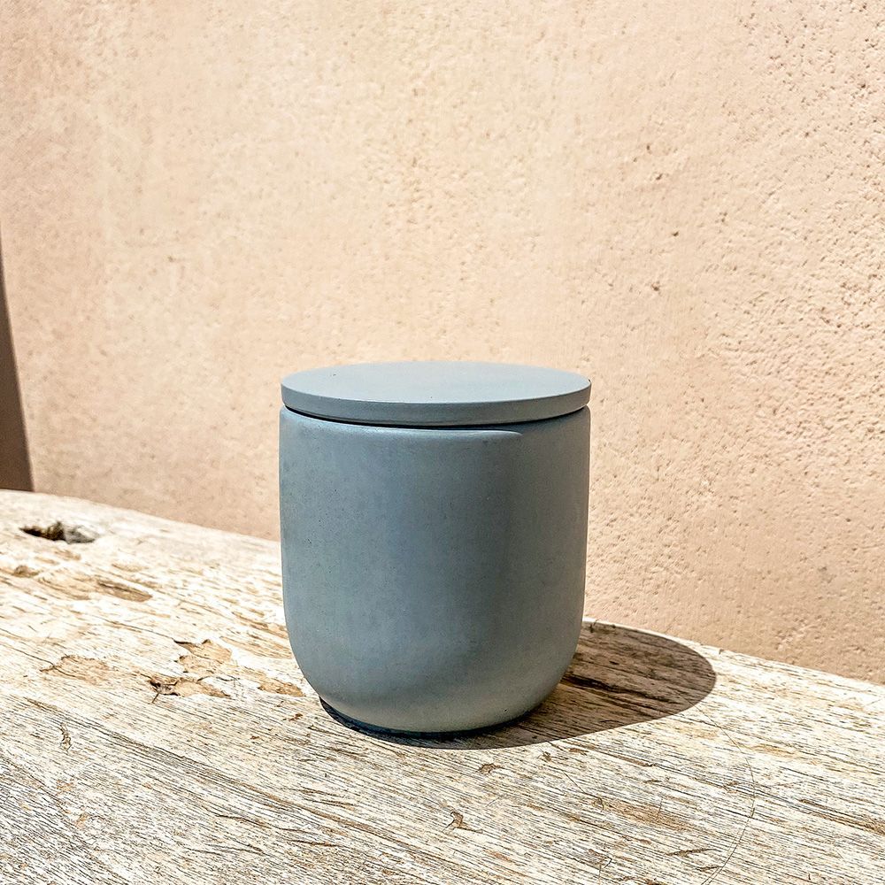 Bougie bleue allumée dans un récipient rond en céramique, couvercle à côté, sur une surface en bois, avec un mur beige en arrière-plan.