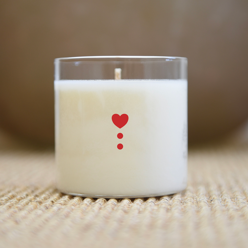 Bougie blanche dans un pot en verre transparent avec un cœur rouge et deux points ; sur une surface tissée.