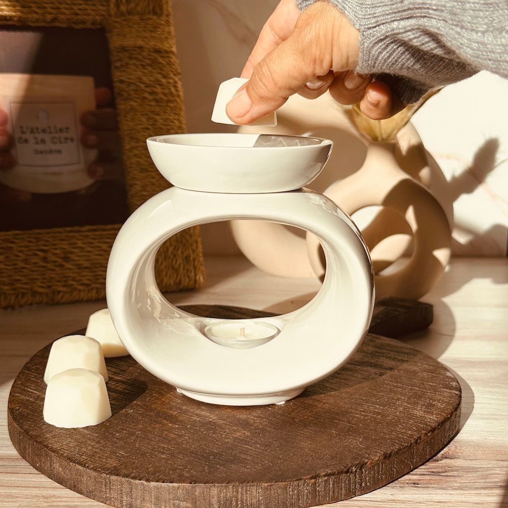 Personne déposant de la cire fondue dans un réchaud en céramique blanche. Base en bois marron.