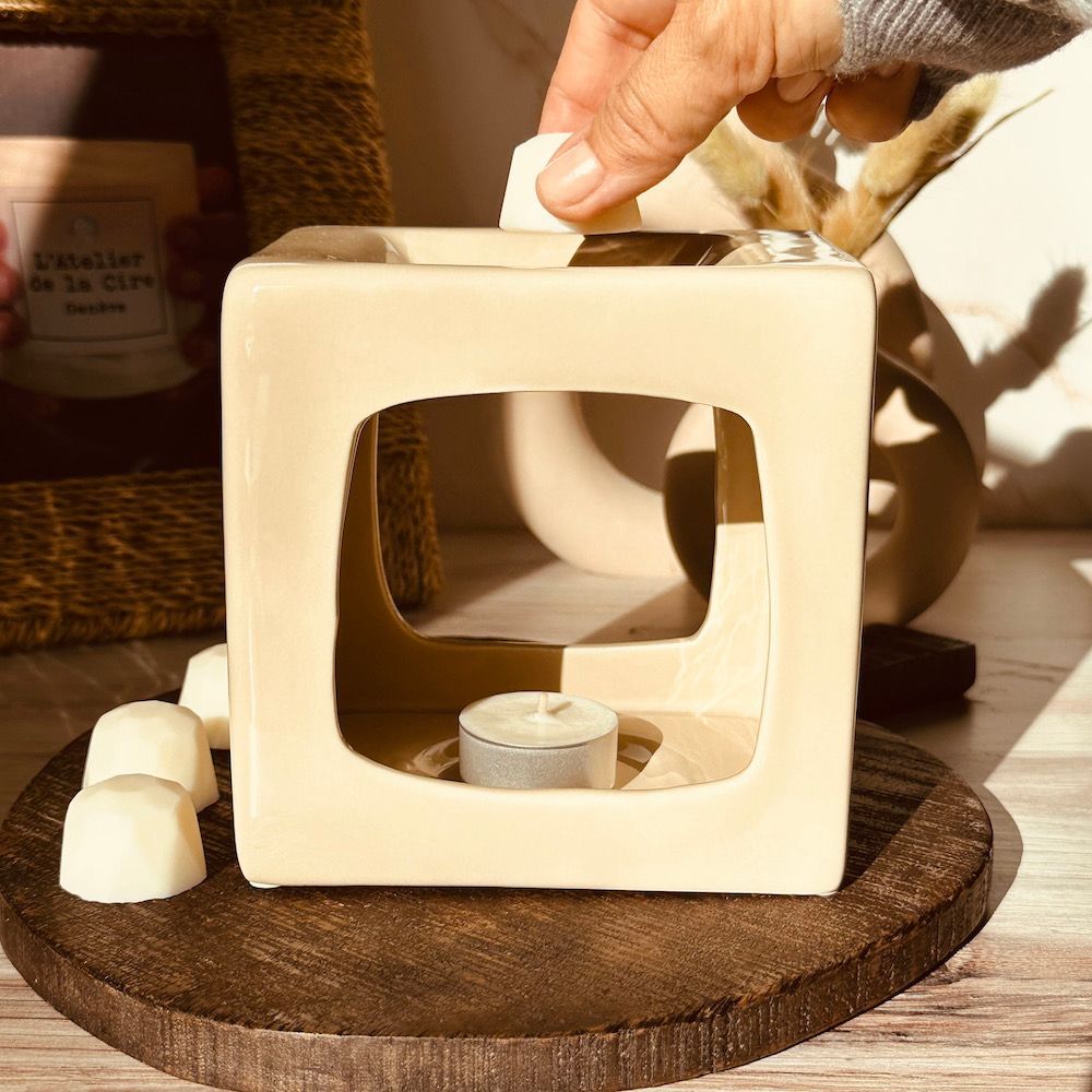 Une personne place de la cire fondue dans un réchaud en forme de cube beige sur une planche de bois.
