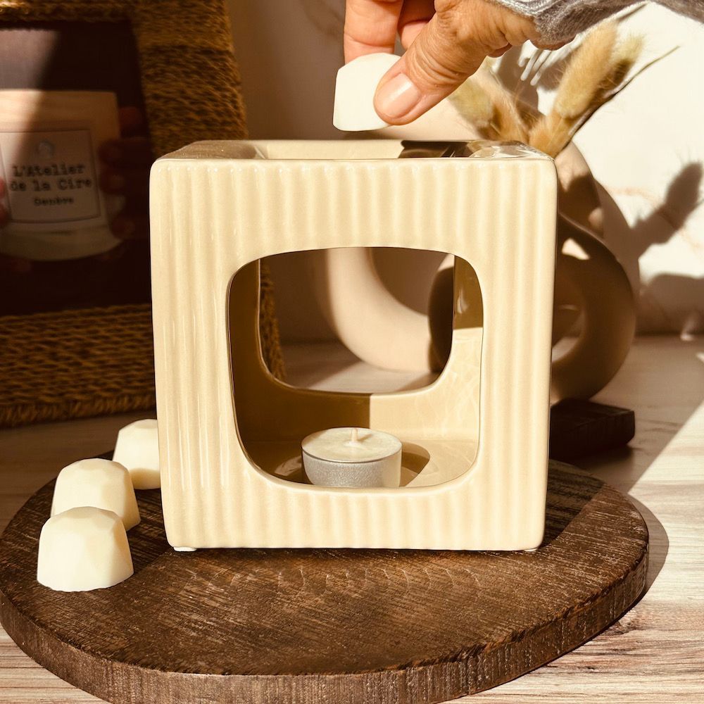 Une main dépose une cire fondante dans un chauffe-cire en céramique beige. Une bougie chauffe-plat est allumée à l'intérieur.