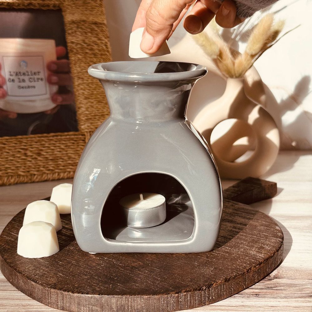 Ajout manuel d'une cire fondue dans un chauffe-plat en céramique grise avec une bougie chauffe-plat allumée, sur un plateau en bois.