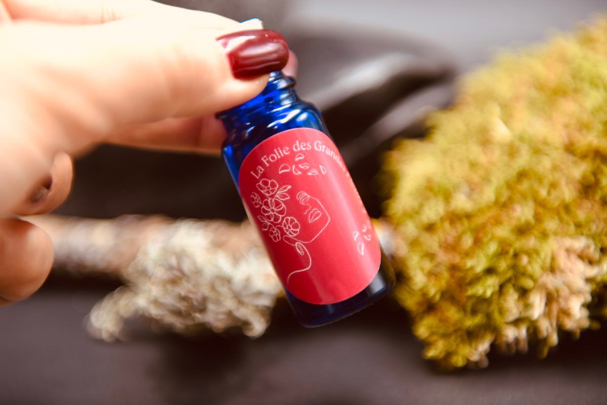 Hand holding a small blue bottle with a red label; blurred moss in the background.