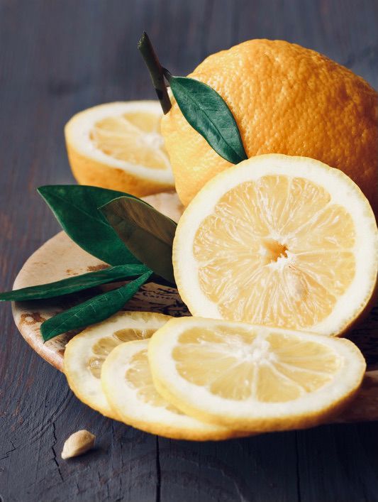 Lemons, whole and sliced, on a wooden plate, with green leaves, against a dark wooden background.