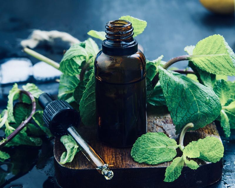 Brown bottle of essential oil with dropper, surrounded by fresh mint leaves on a wooden board.