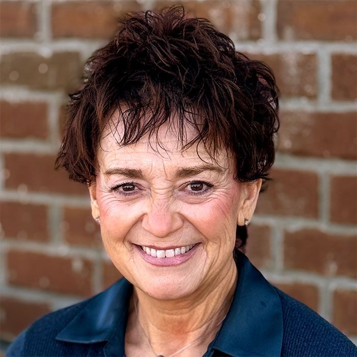 Woman with short, dark, curly hair smiles broadly against a brick wall background; she wears a dark top.