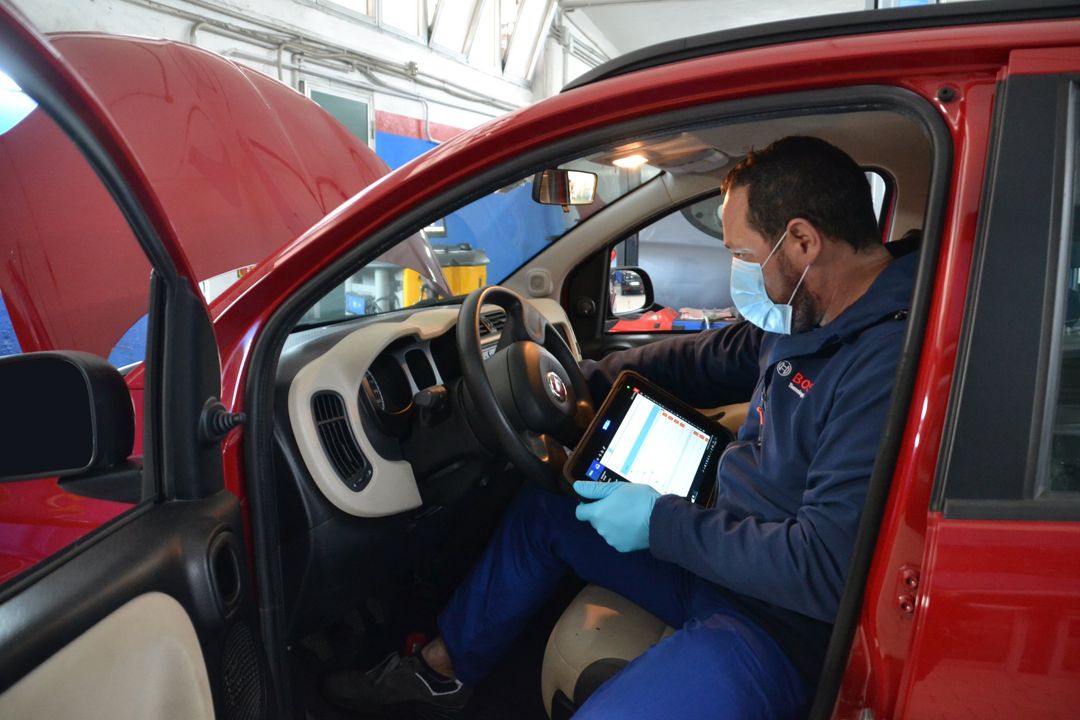Un meccanico a bordo di un'auto rossa usa un tablet mentre è seduto al posto di guida, con il cofano aperto, in un'officina.