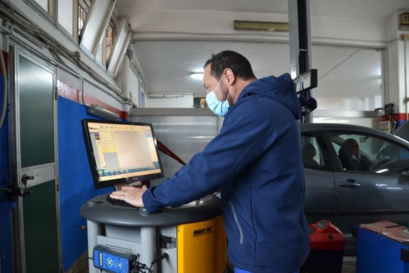 Autofficina e Centro Revisioni Lombardo Giuseppe - Meccanico Palermo usa un computer a una postazione di lavoro in un garage accanto a un'auto parcheggiata.