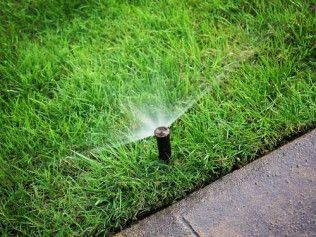A lawn sprinkler sprays water over a green grass field in front of a residential apartment building.