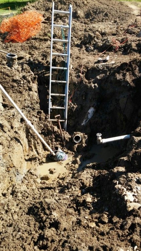 A ladder stands in a muddy, excavated construction trench featuring exposed white PVC pipes.