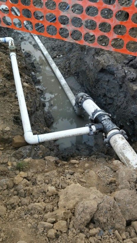 A white PVC pipe assembly with a T-fitting sits in a muddy trench alongside an orange construction safety fence.