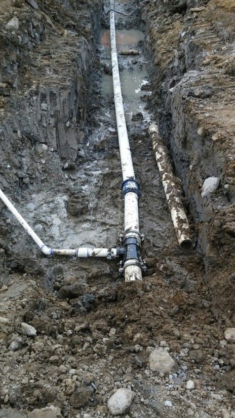 A muddy trench containing white PVC pipes connected by a dark T-joint, with an additional smaller pipe extending sideways.