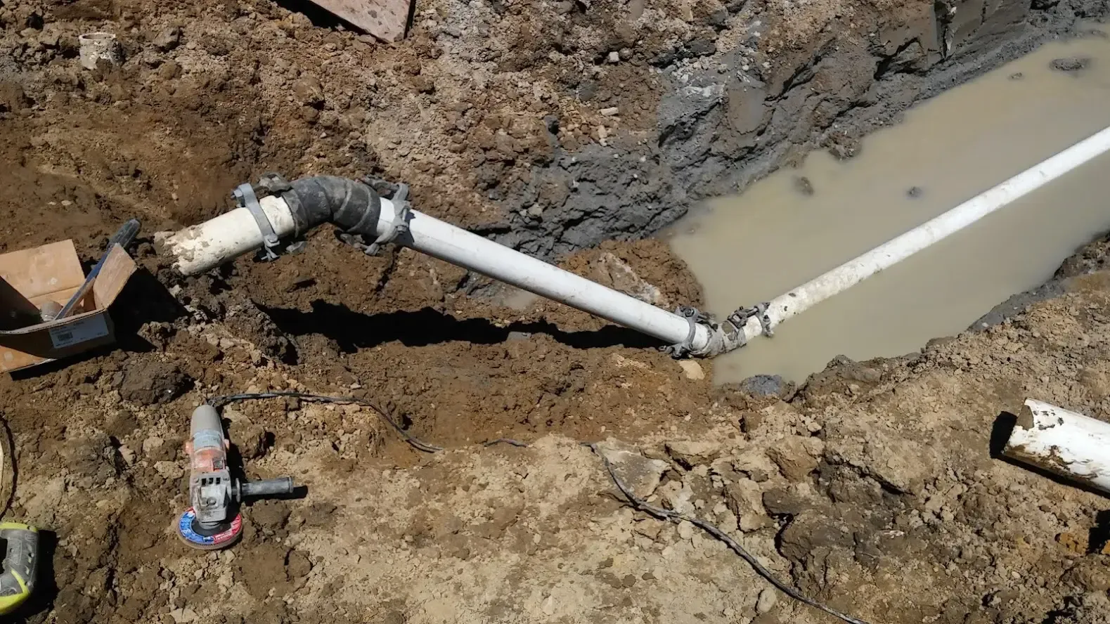 A trench filled with murky water shows white PVC pipes connected by a dark elbow fitting, with a power tool on the dirt.