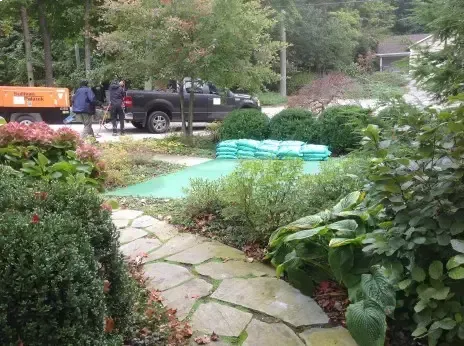 A stone path leads toward a landscape crew working near a truck and a stack of green supplies in a lush garden.