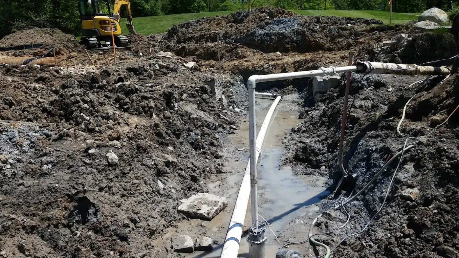 A muddy construction site features an exposed white plumbing pipe and a yellow excavator in the background.
