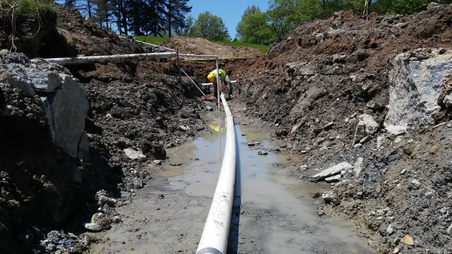 A worker in a high-visibility vest stands in a muddy construction trench next to a long white pipe.