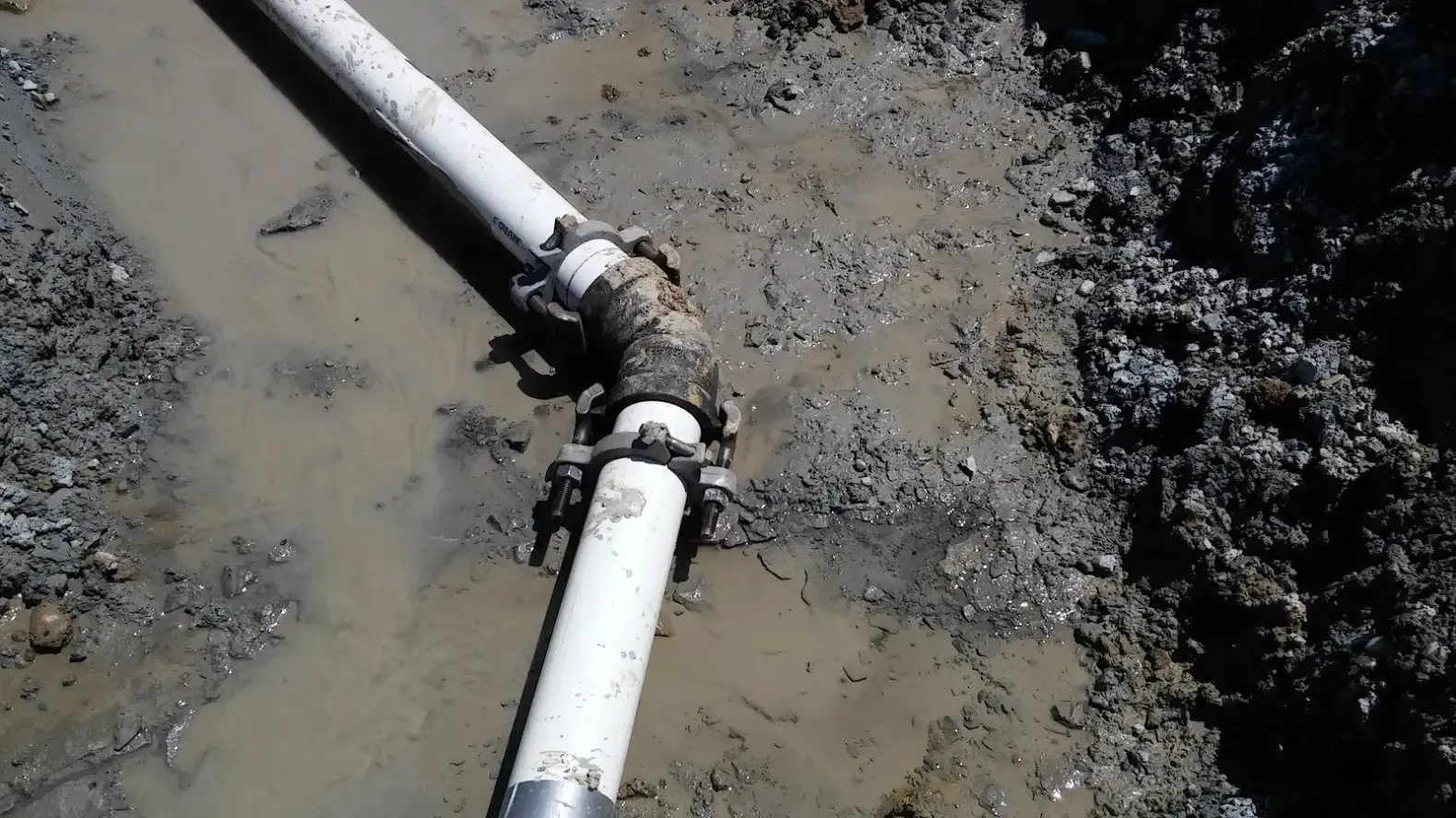A white pipe joint with mechanical couplings, situated in a muddy, excavated trench.