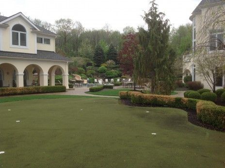 A putting green sits in the courtyard between two houses, surrounded by manicured hedges, trees, and landscaping.