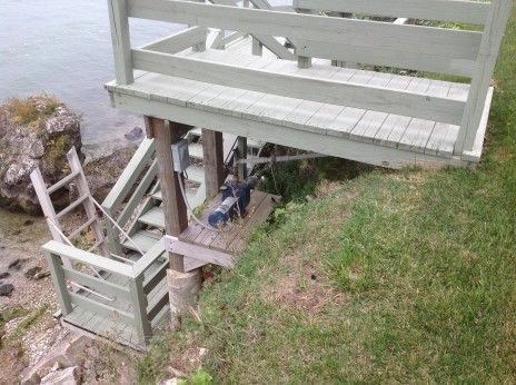 A wooden deck platform and staircase overlook a rocky shoreline, featuring a mounted pump motor on a small lower shelf.
