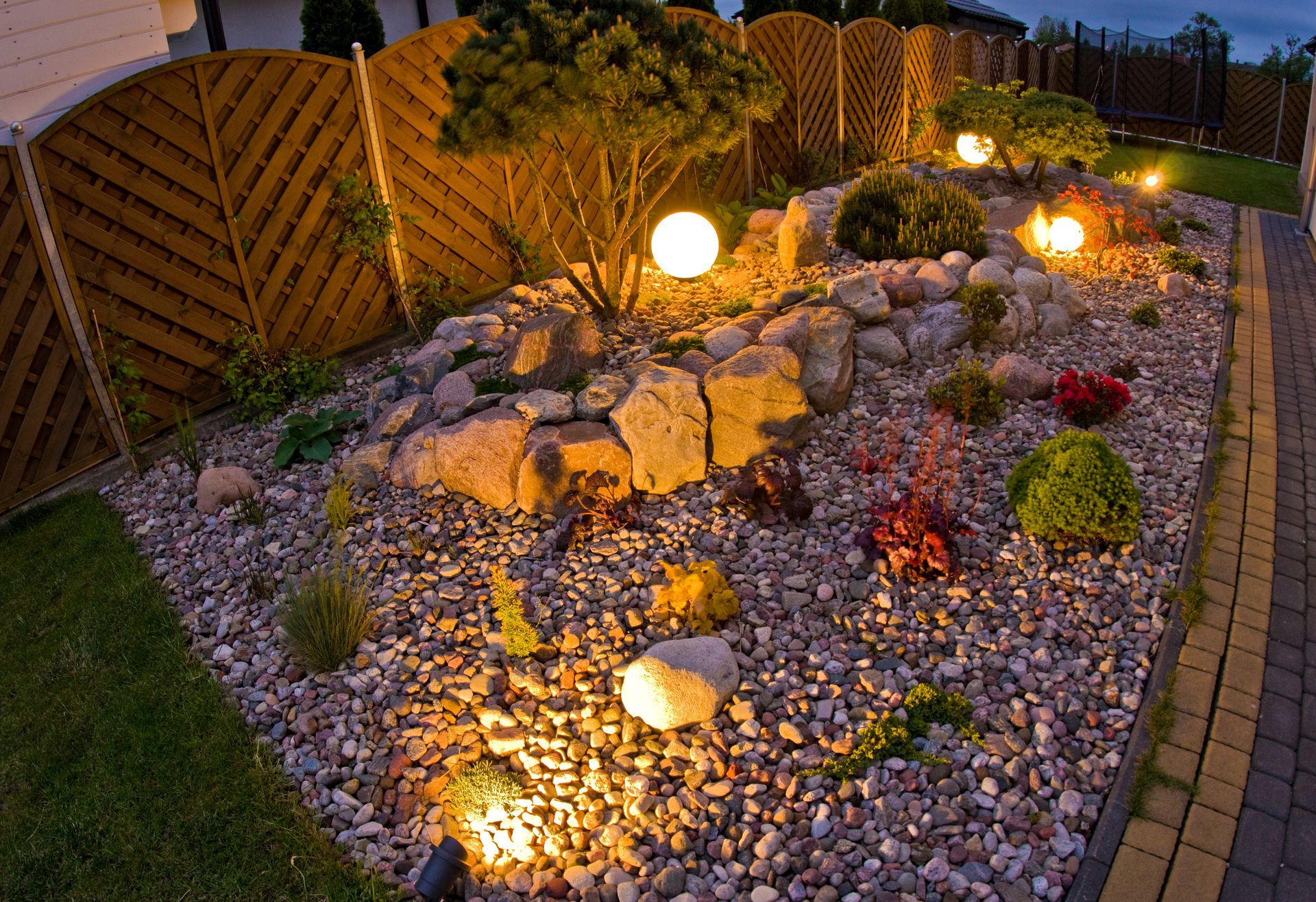 A rock garden at night with glowing spherical lanterns, small shrubs, and a decorative wooden fence backdrop.