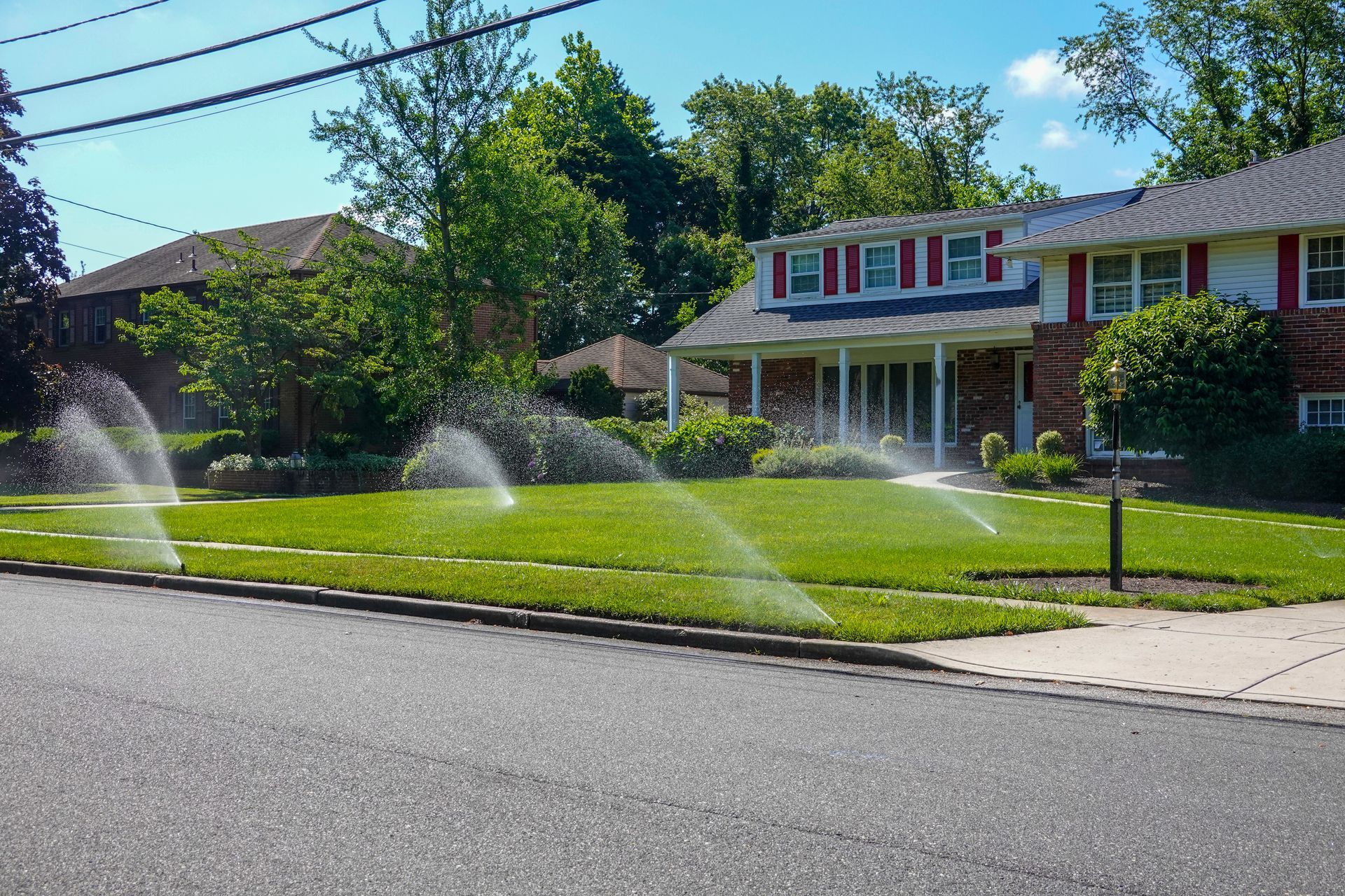 Lawn sprinklers spraying water across a green front yard in a sunny residential neighborhood.