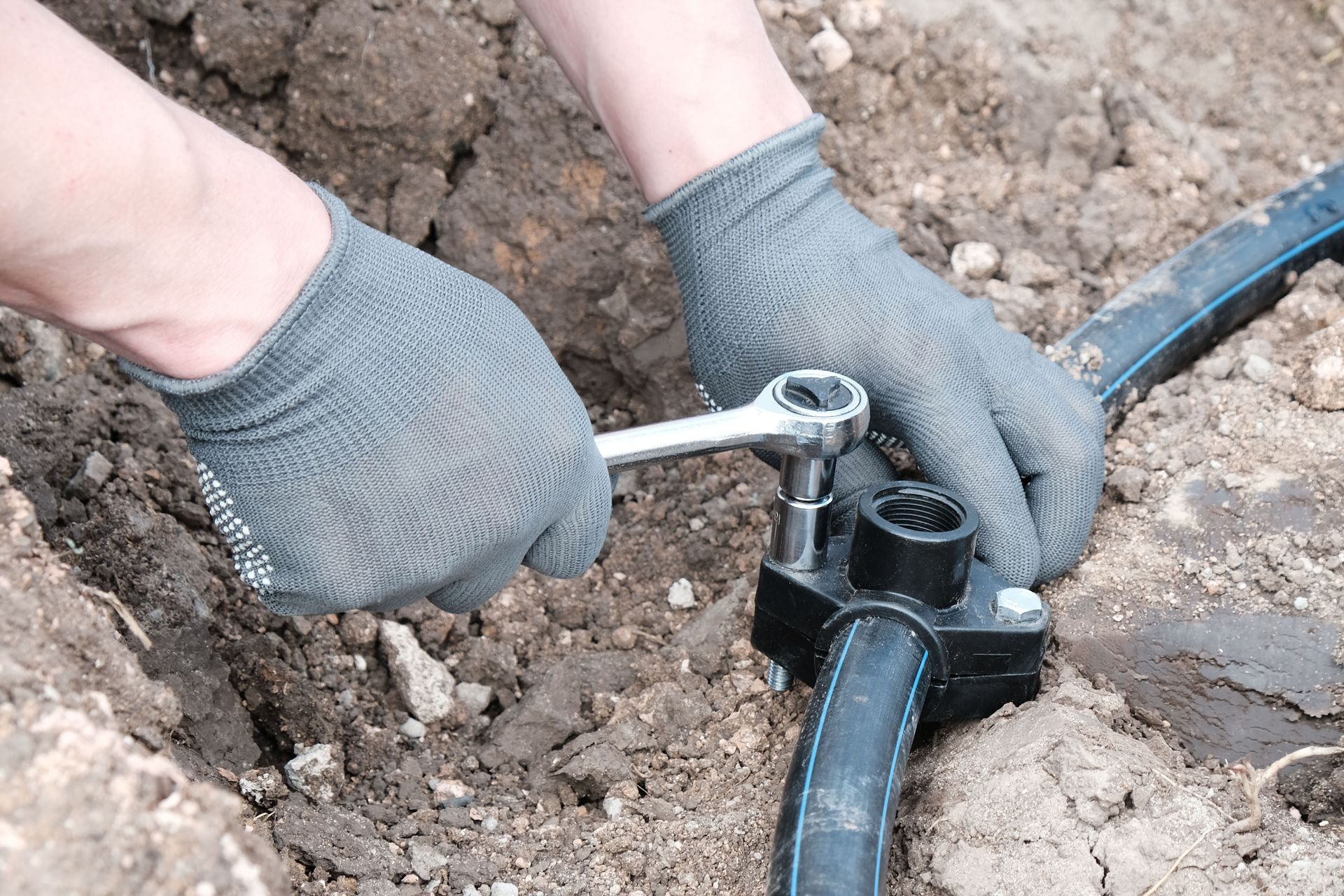 Hands in grey work gloves use a ratchet tool to tighten a clamp on a black irrigation pipe buried in soil.
