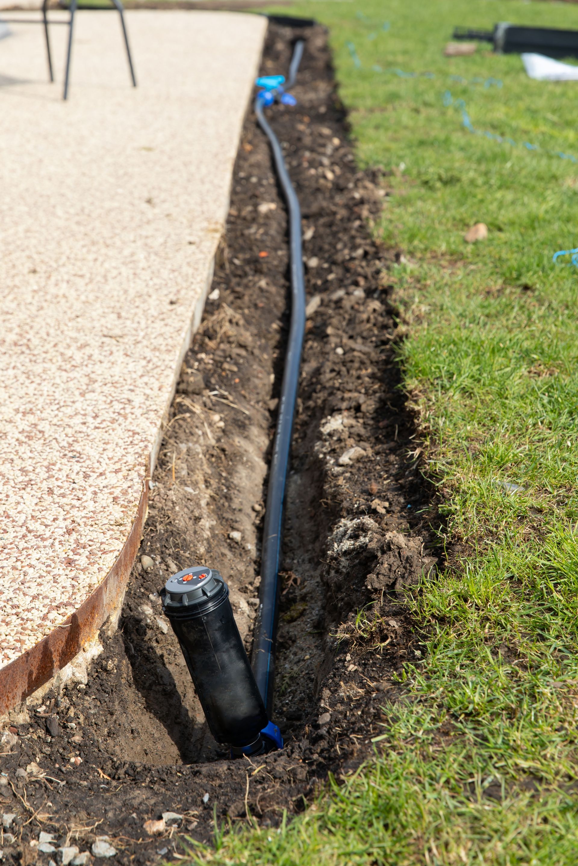 A black irrigation sprinkler head is connected to a plastic supply line in an open trench beside a patio and green lawn.