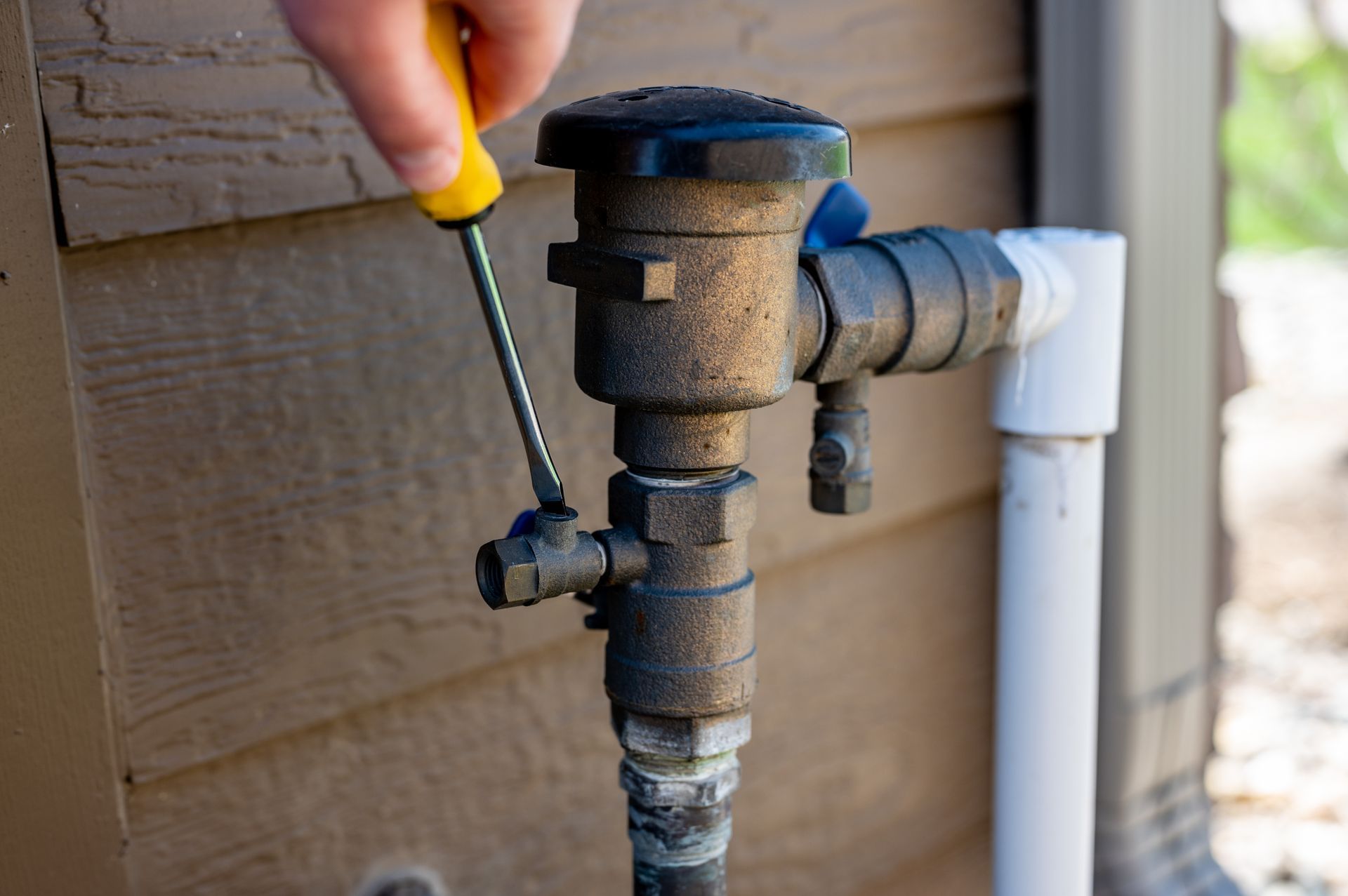 A hand uses a yellow-handled screwdriver to adjust a small valve on an outdoor brass backflow prevention device.