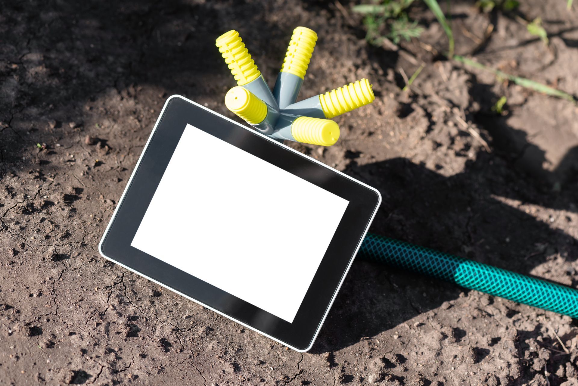 A black tablet with a blank white screen lies on dirt next to a green hose with a multi-directional sprinkler attachment.