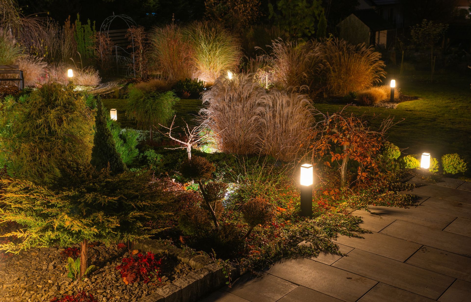 A night view of a landscaped garden featuring several illuminated bollard lights casting a warm glow on plants and a path.