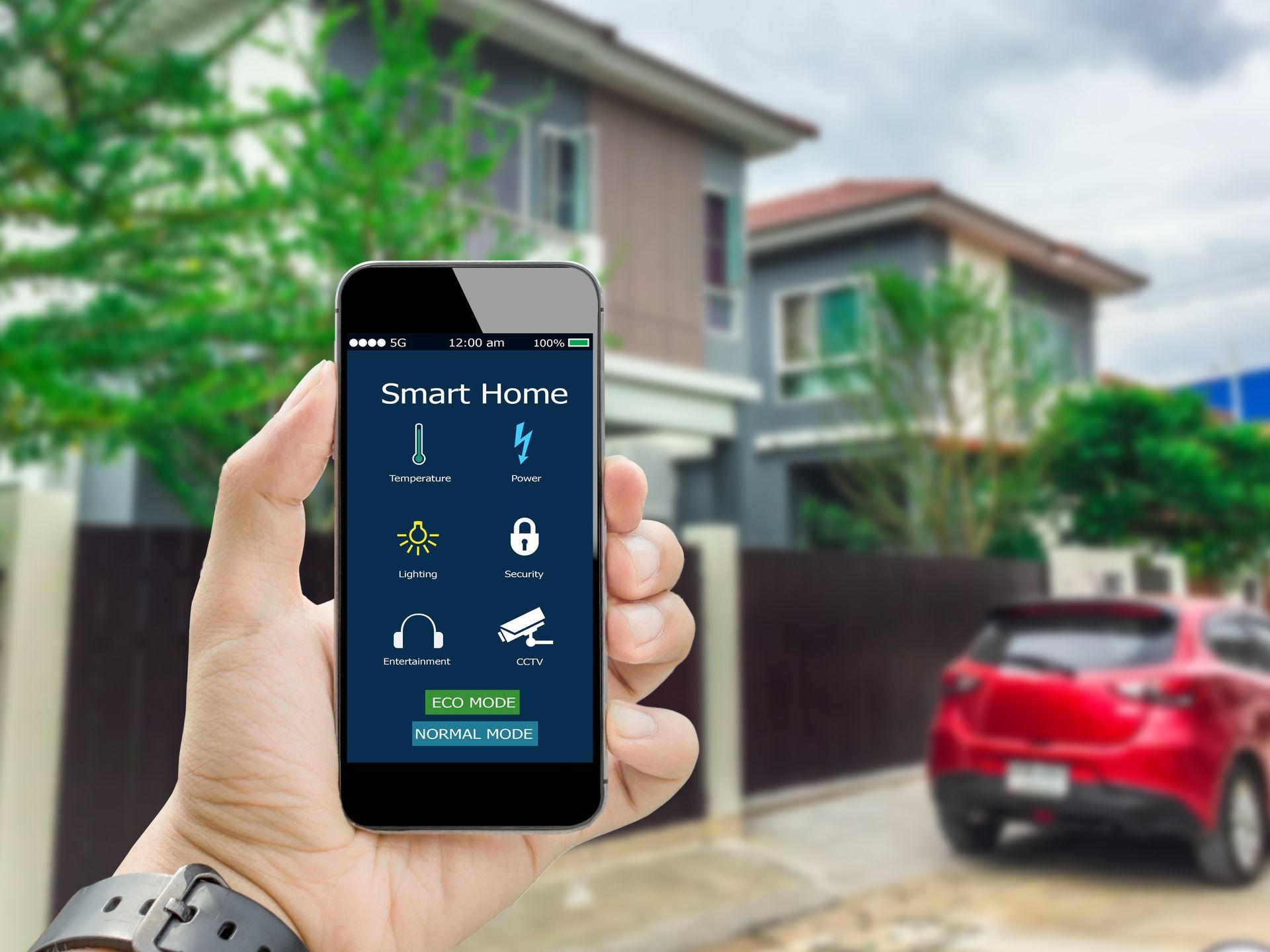 A hand holds a smartphone displaying a smart home control app against a blurred background of a modern house and car.