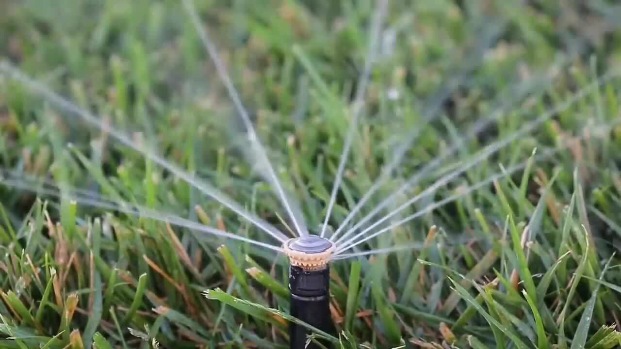 A lawn sprinkler sprays water over a green grass field in front of a residential apartment building.