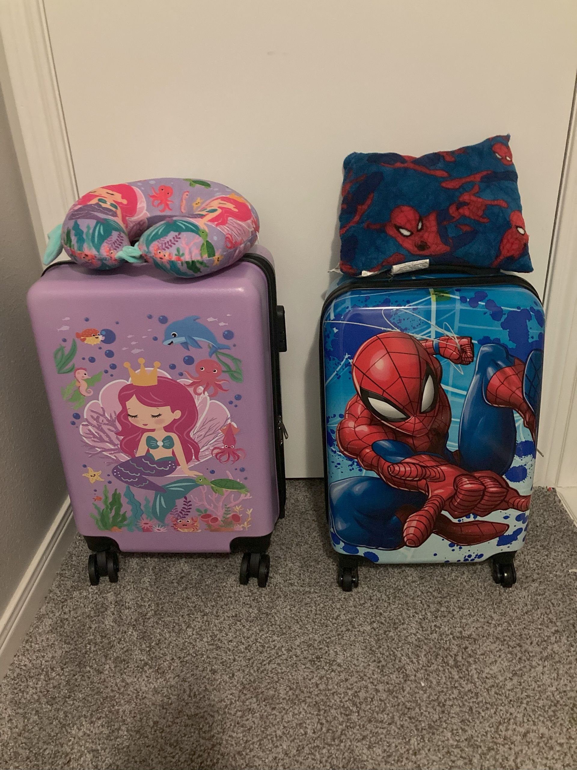 A purple suitcase with a mermaid on it is next to a blue suitcase with a spiderman on it.