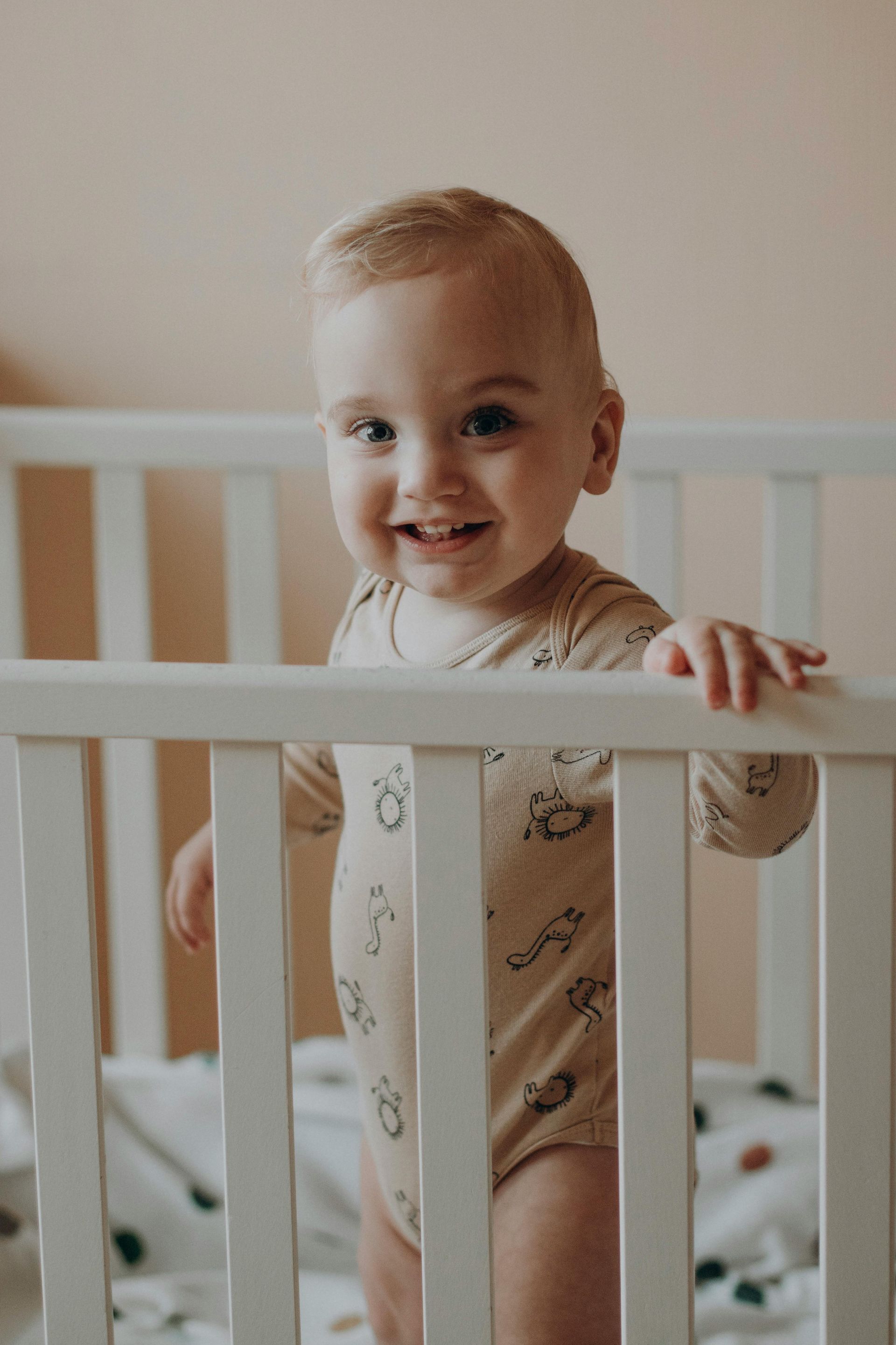 A baby is standing in a white crib and smiling.