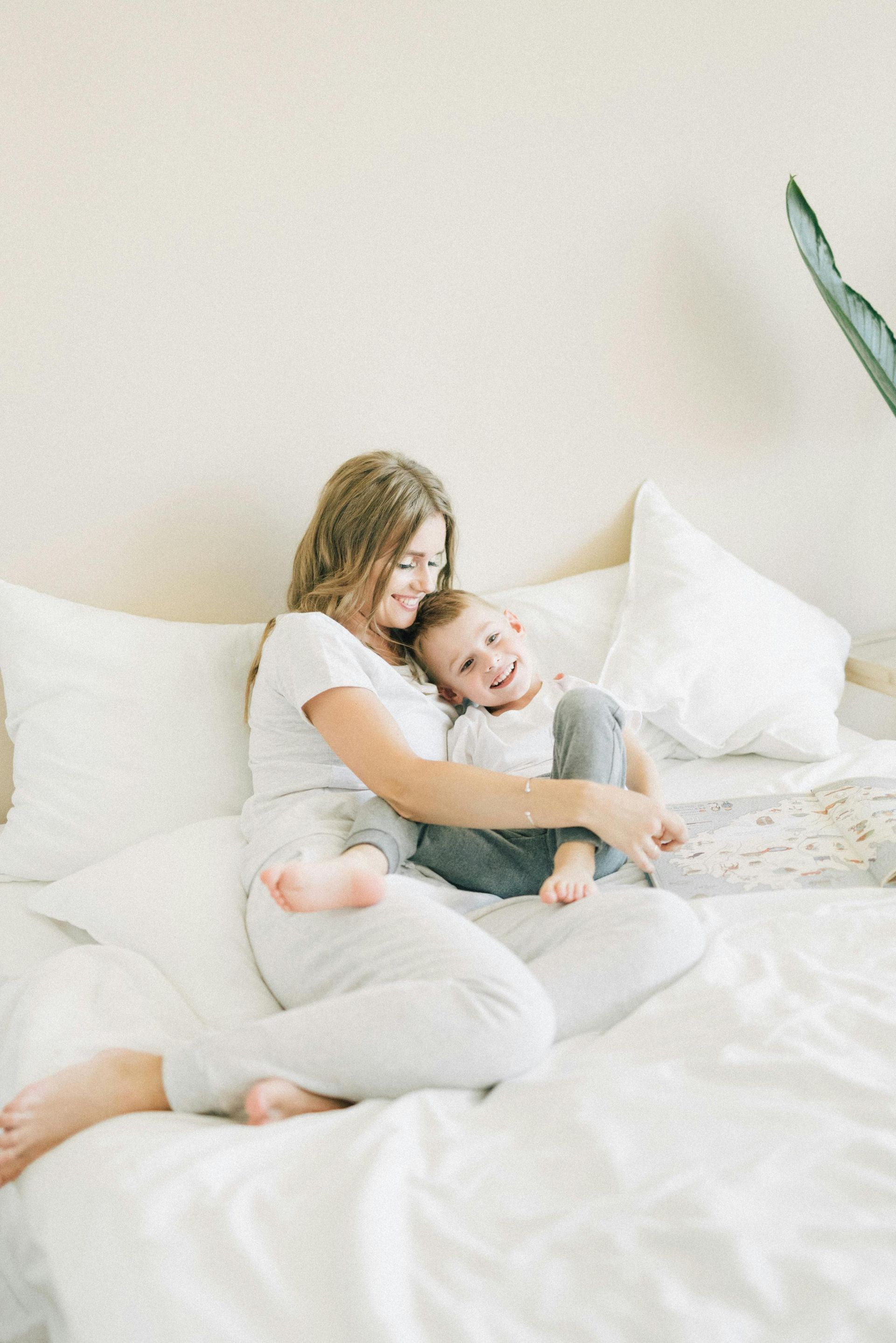 Woman and child laughing on a white bed. Woman in light gray outfit, child in gray pants and white shirt. Bright room.