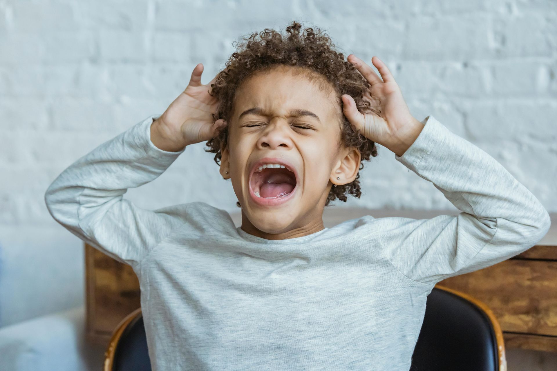 A young boy is sitting in a chair with his mouth open and his hands on his head.