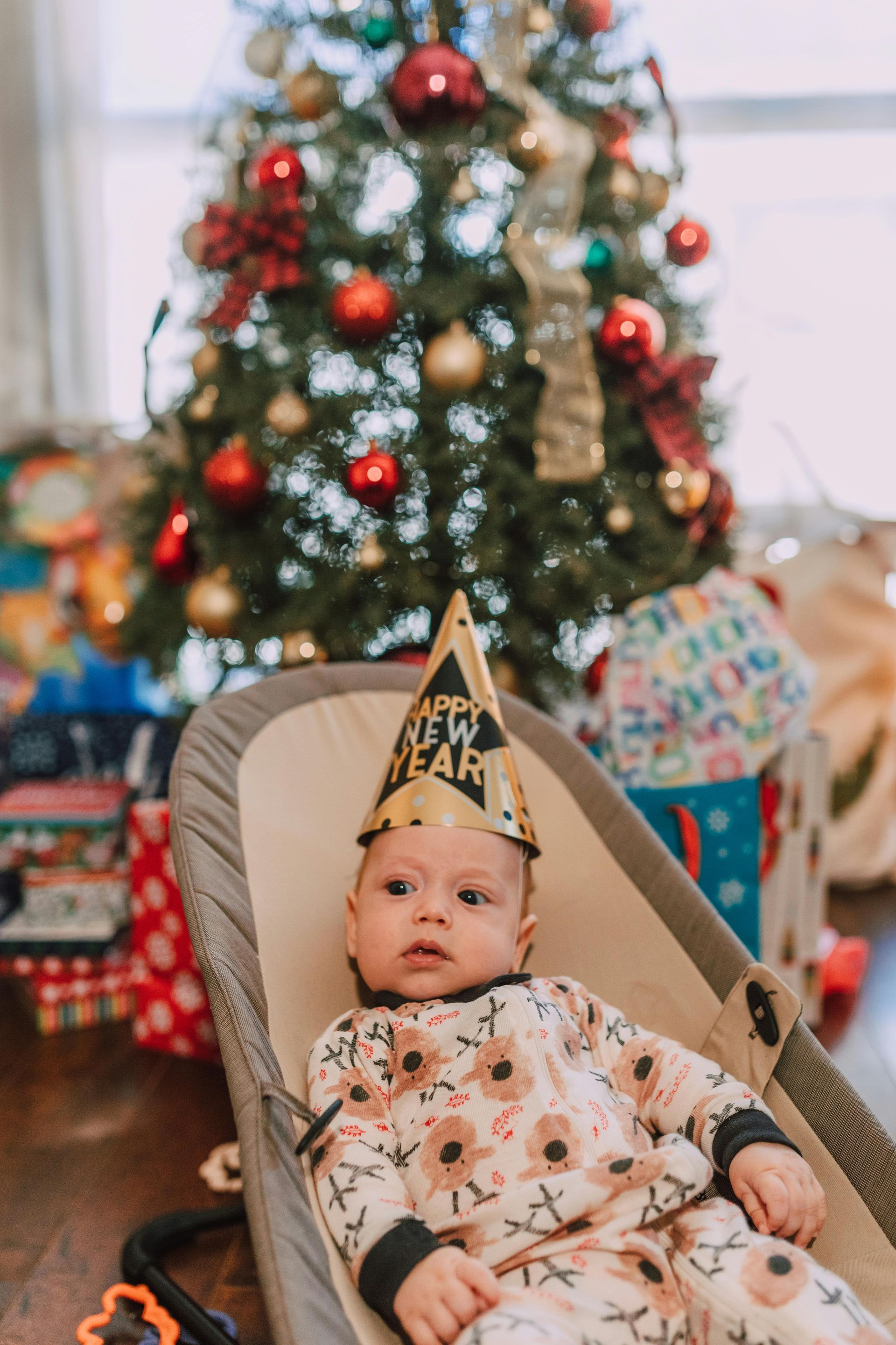 A baby wearing a party hat is sitting in a bouncer in front of a christmas tree.