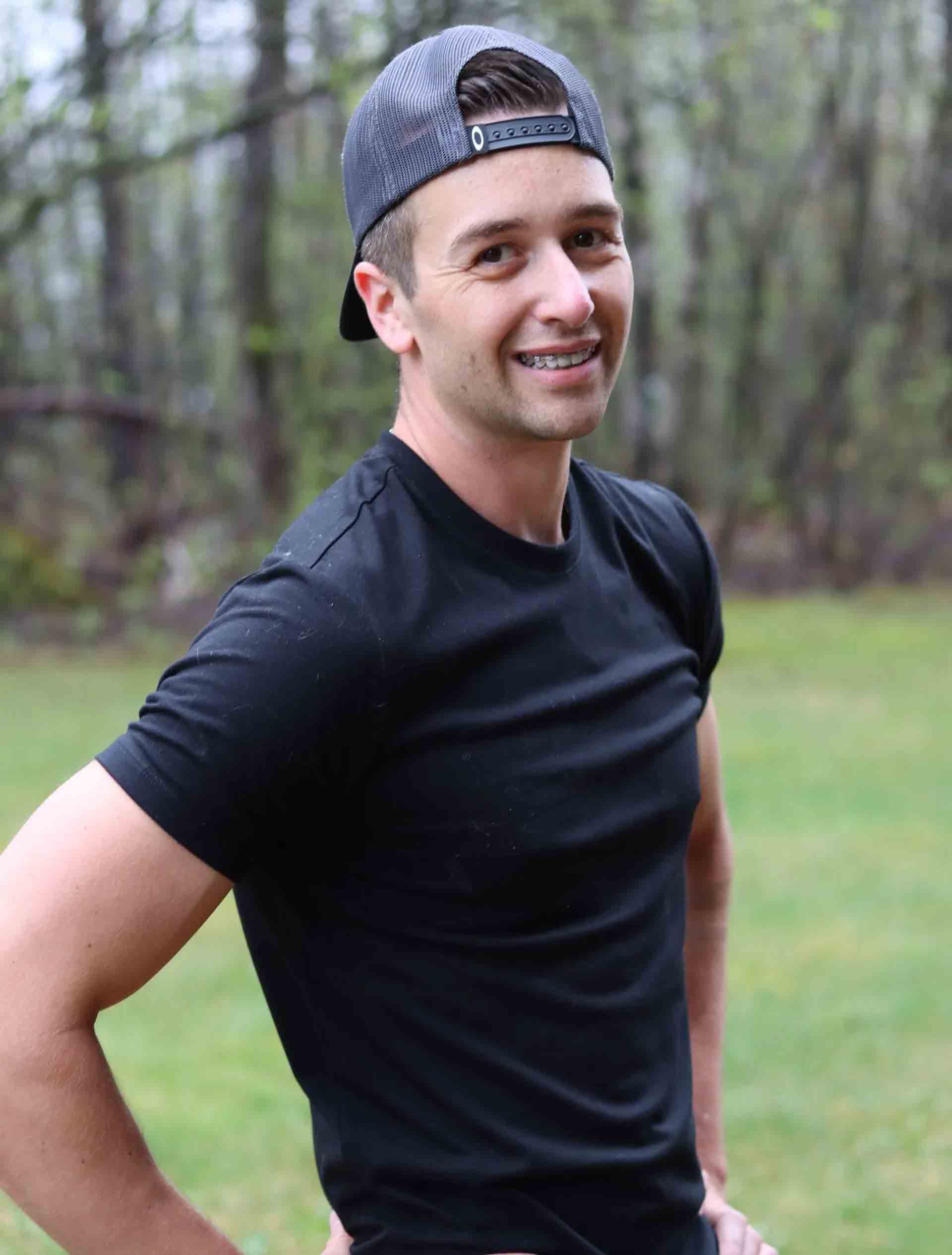 Smiling person in a black T-shirt and backward cap standing outdoors on a grassy path.