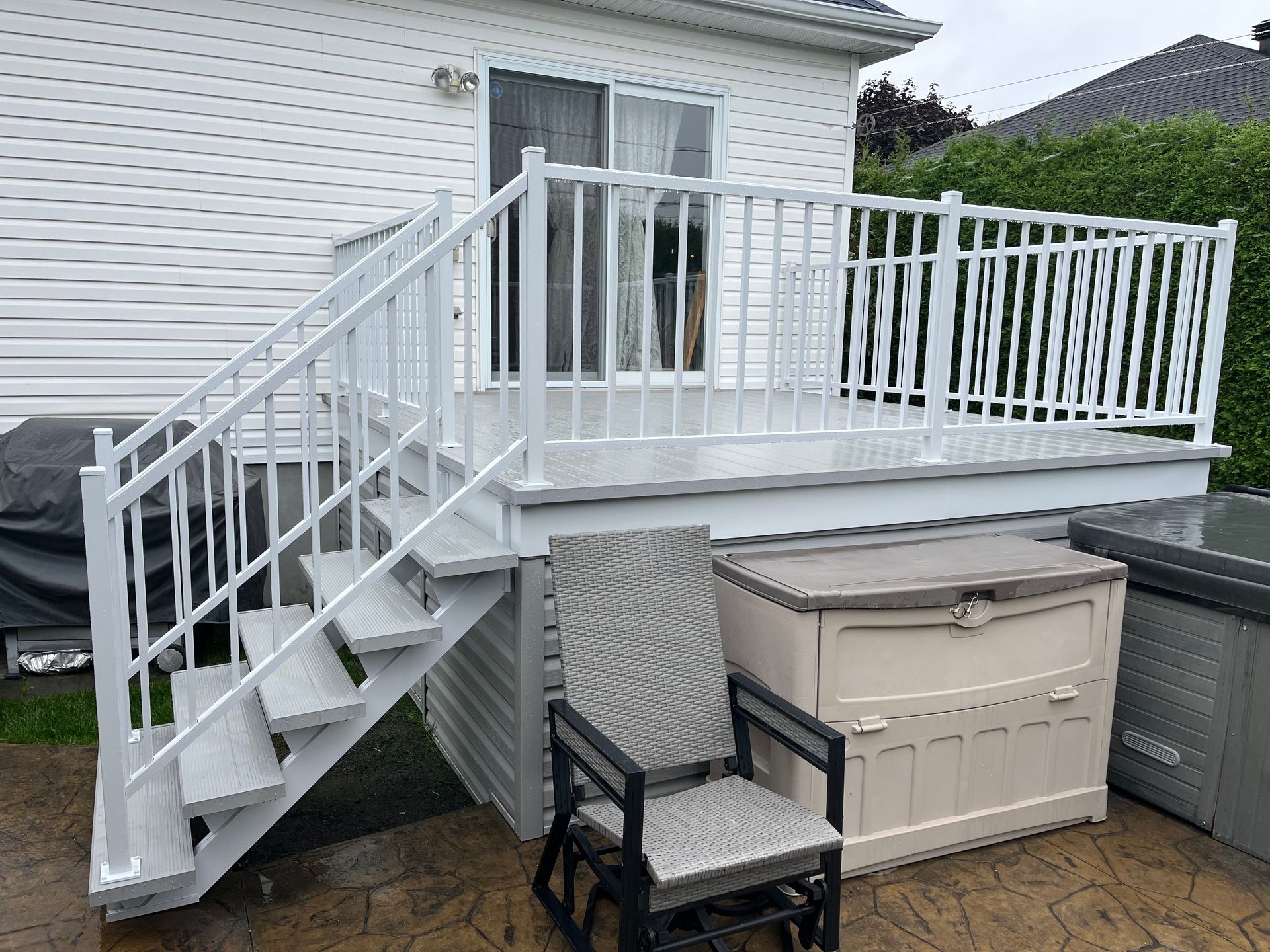 Terrasse blanche avec escalier, rambarde et porte coulissante. On aperçoit une chaise de jardin et un coffre de rangement.