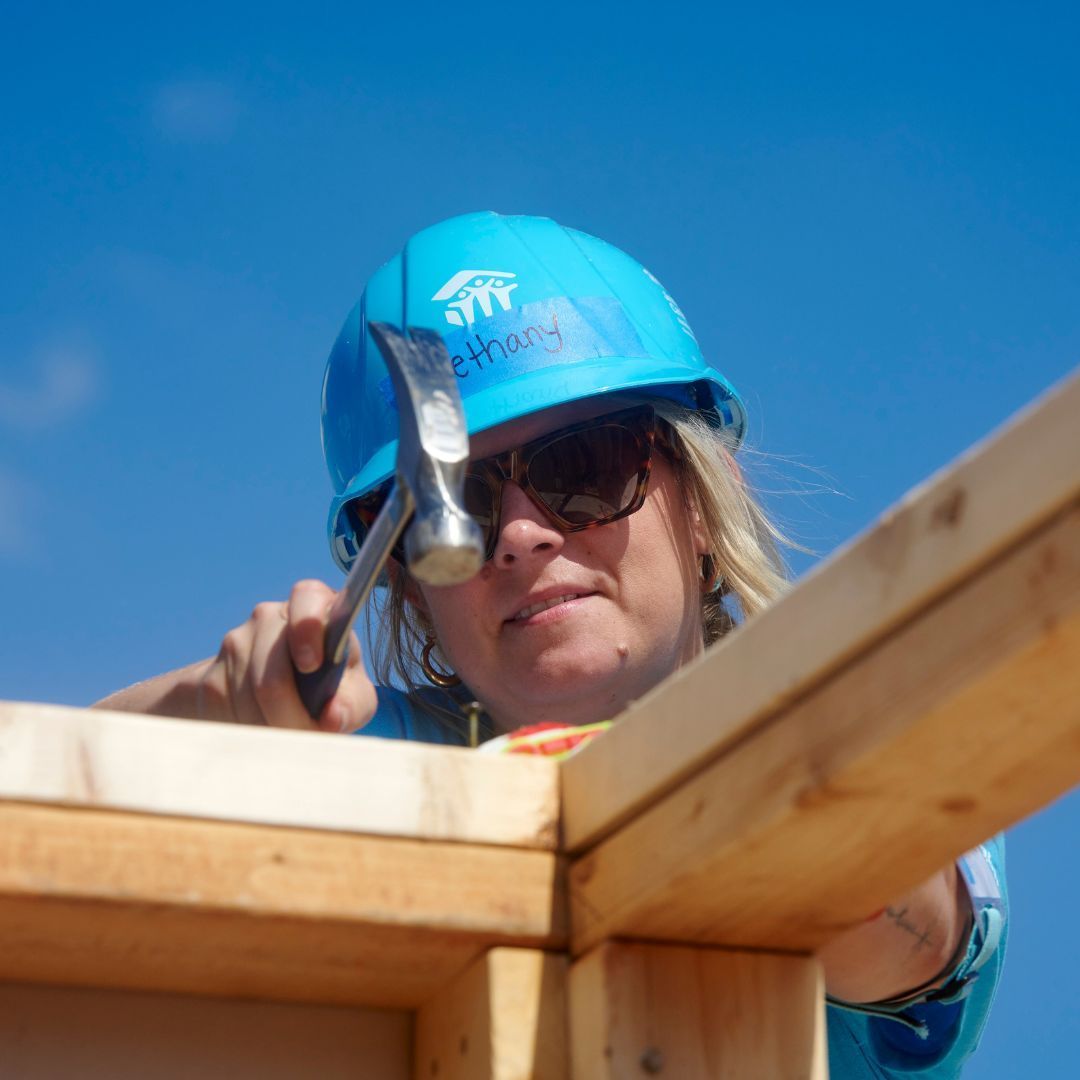 A woman wearing a blue hard hat is hammering a piece of wood