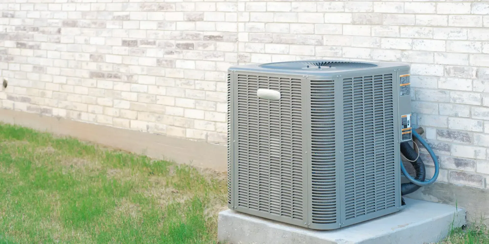 A grey, square residential air conditioning unit sitting on a concrete pad against a brick wall by a patch of grass.