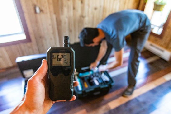 Person holding a black meter displays 625. Another person works on tools in a toolbox, in a room with wood paneling.