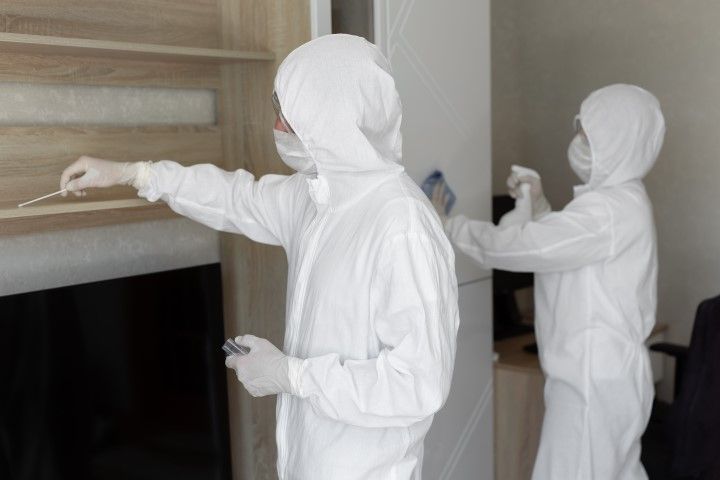 Two people in white protective suits inside, one pointing a tool at a wall.