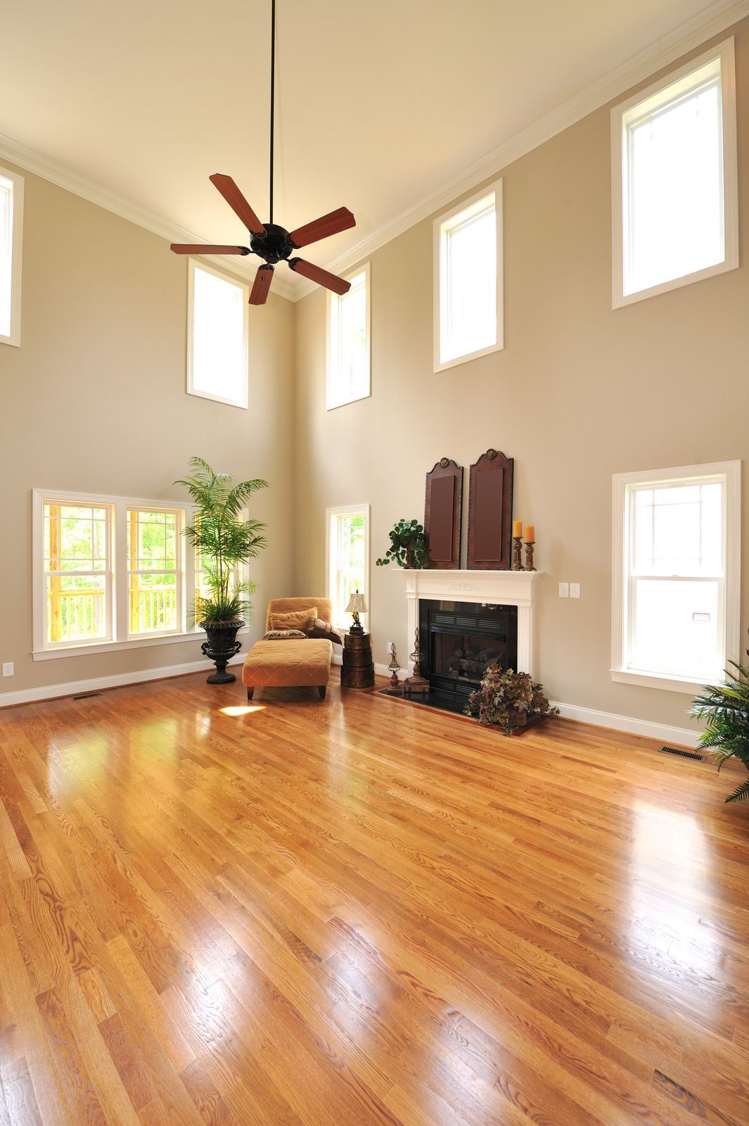 Bright, empty living room with hardwood floors, high windows, ceiling fan, and a white fireplace.