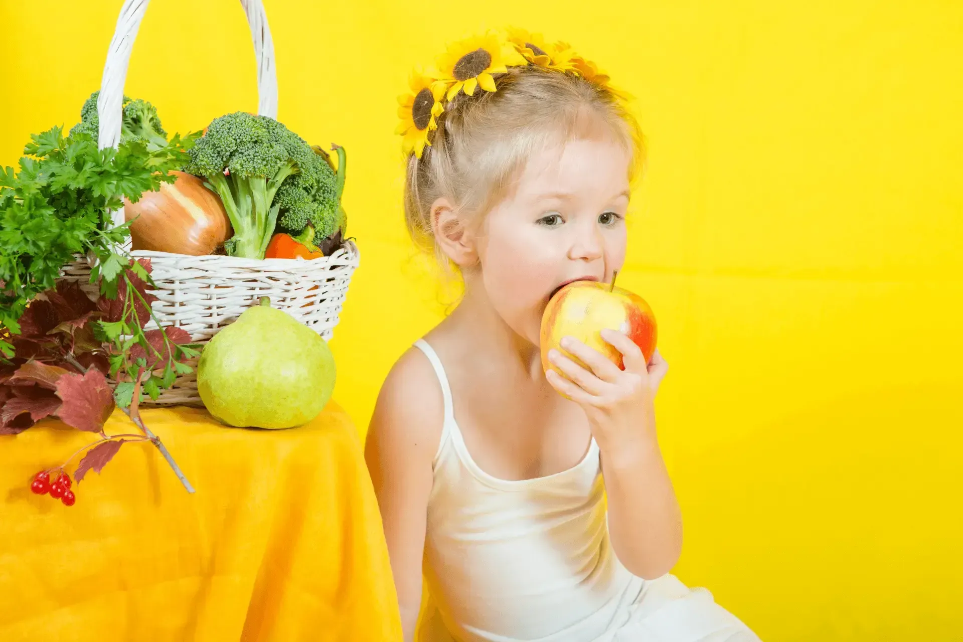 Une petite fille mange une pomme devant un panier de légumes.