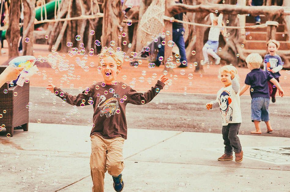 Boys in playground, joyfully reaching for bubbles blown by an unseen person.