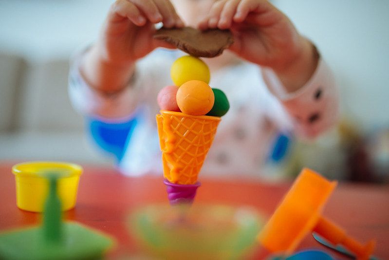 Child making a play-dough ice cream cone with colorful balls and cone on a red table.