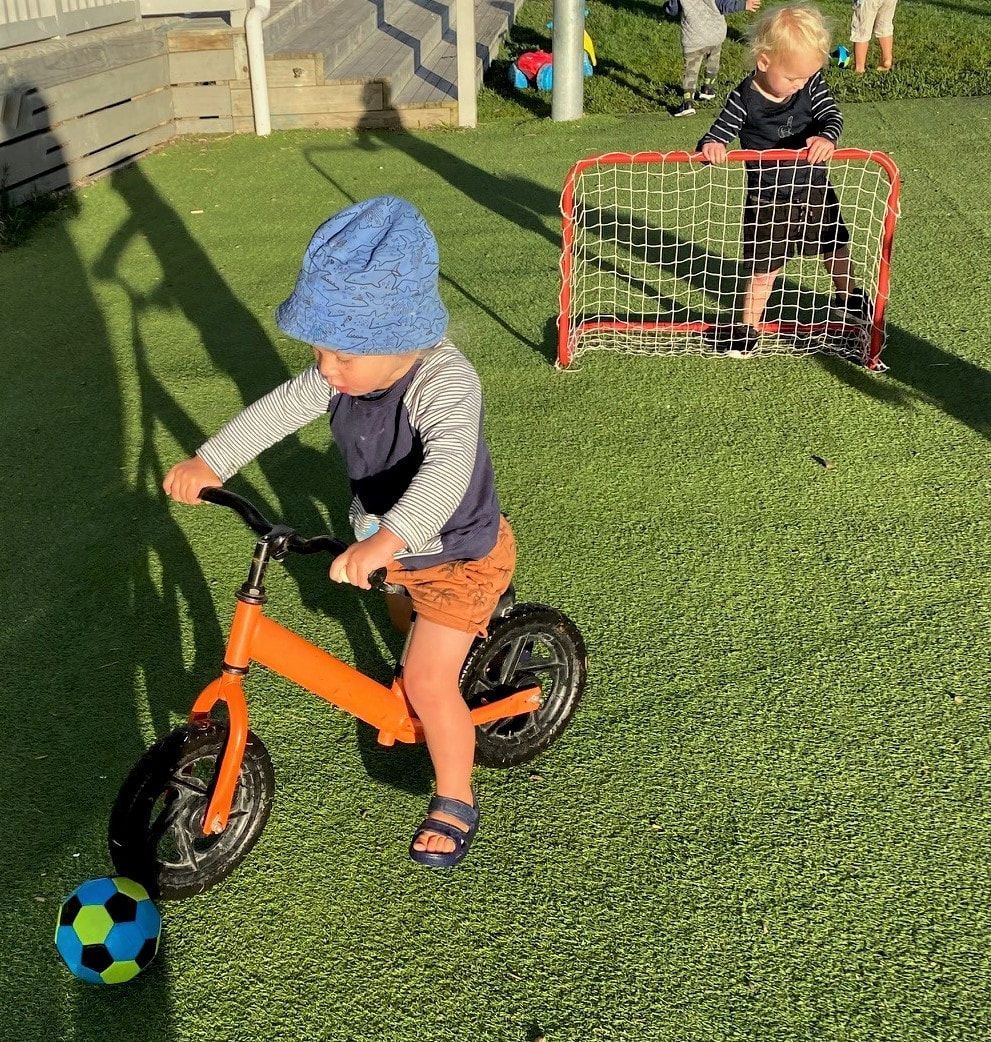 Child on orange balance bike with soccer ball, another child at a small soccer goal on grass.