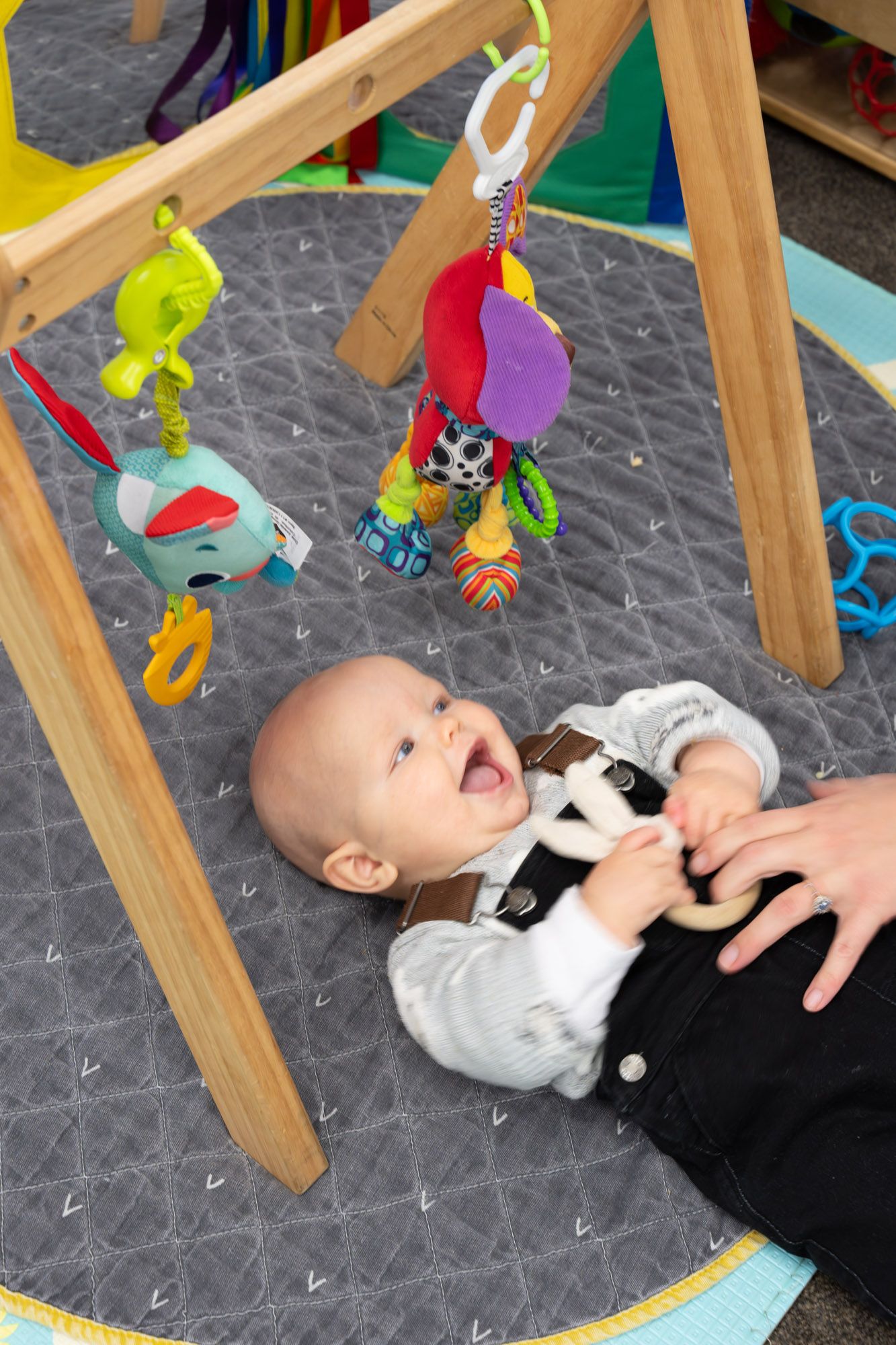 Baby laughing, playing under wooden gym with hanging toys on a gray mat.