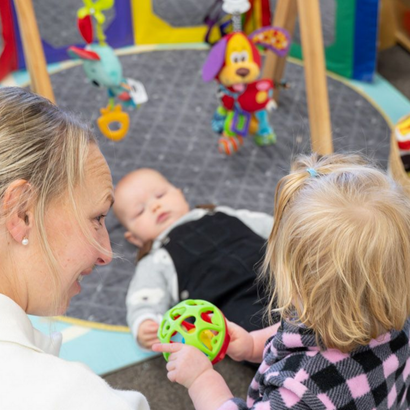 Woman and toddler looking at a toy, with sleeping baby on a playmat. Toys are overhead.